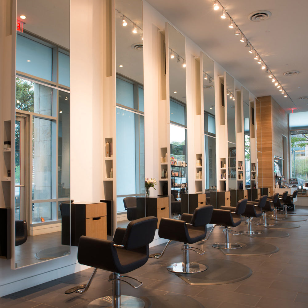 A modern hair salon interior with multiple black swivel styling chairs, wood hair stations, large mirrors, track lighting, and tall windows.