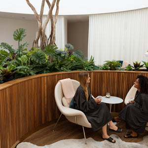 Two women in black robes sit in cream-colored lounge chairs with a small round table between them, inside a modern indoor garden with curved wooden slat walls, lush green plants, and white curtains in the background.