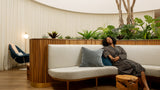 A woman in a gray robe reclines on a curved white sofa with a blue pillow in a modern indoor lounge setting with wood slat paneling, plants, and a tree trunk side table.