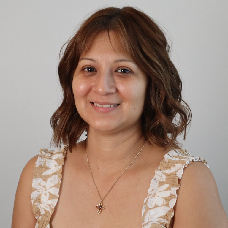 A smiling woman with shoulder-length brown hair wearing a beige and white floral-patterned ruffled top and a gold cross pendant necklace, photographed against a plain light gray background.
