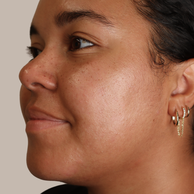 Close-up side profile of a woman wearing small gold hoop and chain earrings against a plain warm beige background.