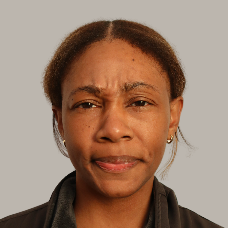 A close-up portrait of a woman with dark brown hair pulled back, wearing a dark brown jacket against a solid light gray background.