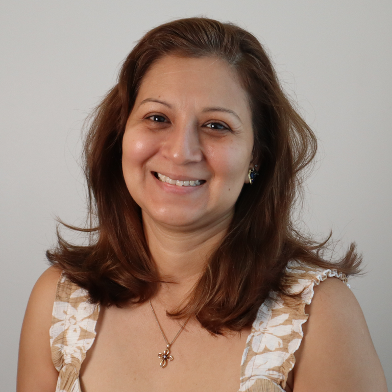 A smiling woman with shoulder-length brown hair wearing a beige and white floral-patterned ruffled top, a gold cross necklace, and a small earring, photographed against a plain light gray background.