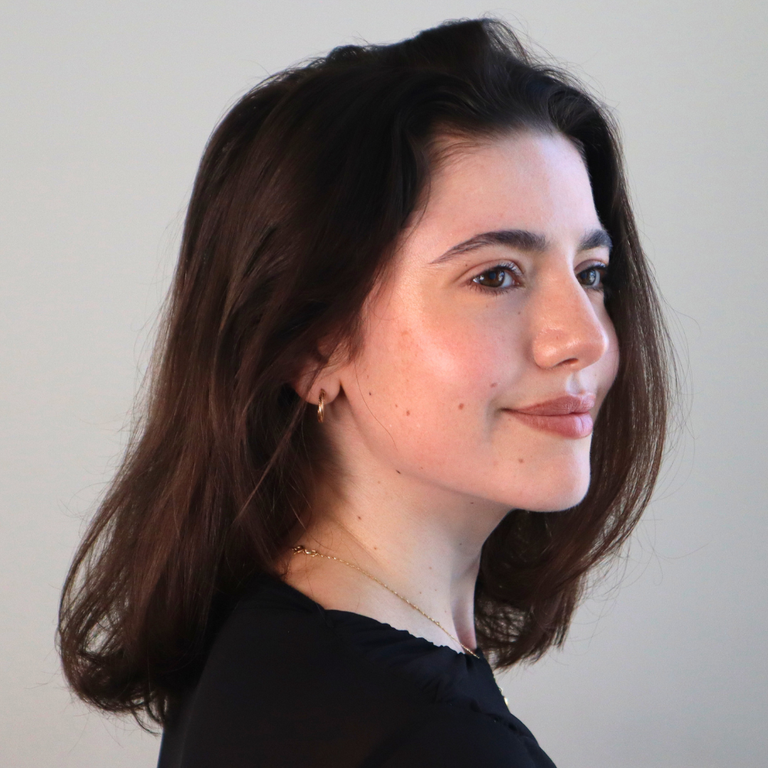 A woman with shoulder-length brown hair is shown in profile against a plain light gray background, wearing a black top and a delicate gold necklace.