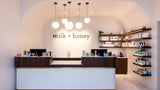 A modern retail counter and shelving inside a boutique with the words 'milk + honey' displayed on the back wall. The space features a white front counter with dark wood vertical slats, multiple spherical pendant lights, and open wall shelves stocked with products.