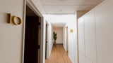 A modern hallway with light wood floors, white walls, recessed ceiling lights, a small potted plant in the distance, and gold room-number wall decor.