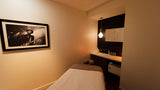 A spa treatment room featuring a massage table in the foreground, a black-framed black-and-white photograph on the beige wall, and a modern vanity area with a hanging pendant light in the background.