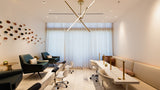 A modern, bright spa or salon interior with teal lounge chairs, white rolling office chairs, a long cream-colored reception desk with gold accents, and a large window covered in sheer white curtains.