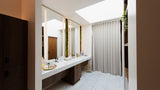 A modern bathroom interior with a long white double vanity, large mirrors, light wood cabinetry, white herringbone tile flooring, and a white pleated shower curtain under a skylight.