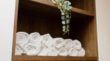 A set of neatly rolled white towels displayed on a wooden shelf, with a trailing green plant hanging over the top shelf in a clean indoor setting.