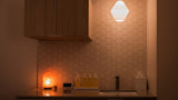 A modern bathroom vanity area with a hexagonal tiled backsplash, wooden cabinets, a countertop sink, and a glowing geometric pendant light above.