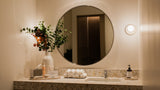 A warm, modern bathroom vanity setup featuring a large round wall mirror, a speckled stone countertop with a sink, a white vase of greenery and orange flowers, a tray of white bath towels, and a soap dispenser.