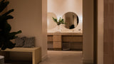 A modern bathroom interior with a double vanity, round mirrors, and warm ambient lighting, viewed from an adjacent hallway with a bench and plant in the foreground.
