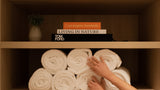 A pair of hands arranging white rolled towels on a wooden shelf, with books and a small vase displayed on the upper shelf.