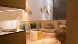A modern living room corner featuring a beige upholstered corner sofa with multiple accent pillows, a white and yellow cylindrical ottoman, a white cowhide rug, and warm ambient lighting.