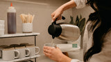 A person pouring hot water from a stainless steel kettle with a black lid into a white mug on a shelf in a kitchen or cafe setting.