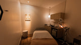 A compact spa or massage room with a single treatment bed, warm ambient lighting, wood flooring, wall cabinets, and a small vanity area with a fan and mirror.