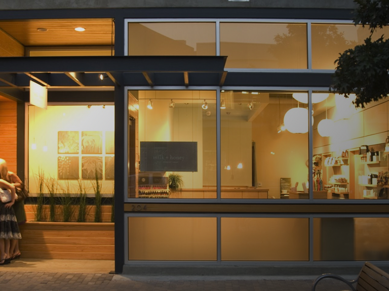 Exterior storefront of a modern building with large glass windows, warm yellow interior lighting, and a person standing partially visible on the left sidewalk.