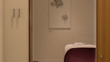 A view through a doorway into a spa or treatment room, showing a massage table with a deep purple throw blanket, a small wooden stool, and a framed abstract black line drawing on the wall.