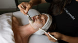 Person receiving a facial treatment with a towel on their head and a brush applying product.