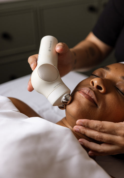 A close-up of a person receiving a facial treatment while another person uses a white handheld device with a metallic roller on their face. The person receiving the treatment is lying down on a white-covered bed or table, with a neutral gray background in the distance.