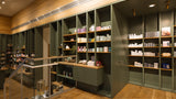 A modern interior retail space with tall built-in shelving in dark green and wood tones, filled with boxed products, set in a warm wood-finished room with a glass railing and ceiling lights.