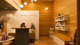 A warm, modern interior space with wood-paneled walls and floor, a round ribbed gray reception desk, white wall-mounted shelves with products, and a built-in bench with cushions against the right wall.