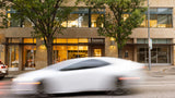 A blurred white sports car drives past a modern office building with storefronts, including a sign reading 'honey' and another reading 'SALON', with trees in front.