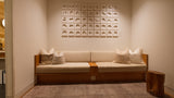 A modern wooden bench with cream cushions and decorative pillows, set against a warm beige wall with a grid of small framed art pieces above. A textured gray rug and a rustic wooden side table are in the foreground.