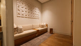 A modern low-profile beige sofa with a wooden base in a minimalist room, decorated with multiple square wall art prints and a gray rug on a wood floor.