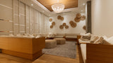 A modern, warm-toned lounge or spa interior with wooden seating, cream cushions, a round glass chandelier, wood ceiling panels, and decorative woven wall pieces.