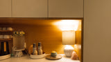 A warm, modern kitchen or bar counter with a built-in wooden shelf illuminated by a glowing lamp. The shelf holds a lit mug, a glass carafe, small bottles, a small plate with a yellow item, and a textured white table lamp with a pink crystal accent.