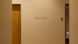 A beige interior hallway wall with a small black sign reading 'TO LOUNGE + LOCKERS' and an arrow pointing left, with a wooden door on the left and an open doorway on the right showing a bed.
