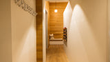 A modern interior hallway with warm wood flooring and walls, a built-in bench with pillows at the end, a hanging pendant light, and a decorative wall-mounted object on the left.