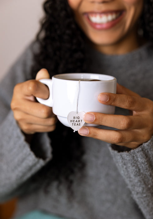 A person in a gray knit sweater holds a white ceramic mug with a tea bag hanging from it. The tea bag tag reads “BIG HEART TEA.