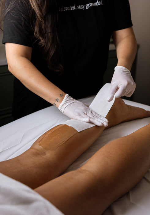 A person lying on a treatment table receives a leg waxing application from a professional stylist wearing a black uniform and white gloves, with white sheets covering the table.