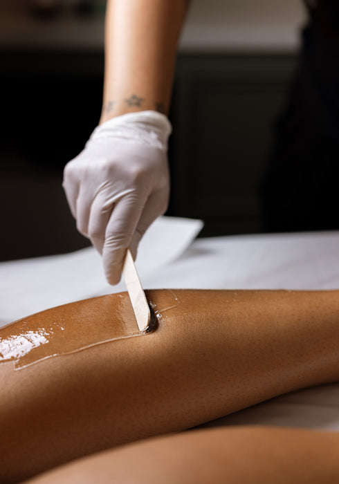 A close-up of a leg being treated with an adhesive hair removal gel, with a gloved hand using a spatula to spread it. The person's leg is resting on a white treatment table.