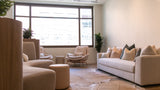 A modern living room featuring a light beige sectional sofa with matching throw pillows, a matching beige armchair, a round ottoman, and a large cowhide rug on a wood floor, with a large window in the background.