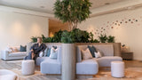 A woman in a dark robe sits on a light gray modular lounge sofa in a bright, modern spa-like interior with wood floors, potted greenery, and matching poufs.