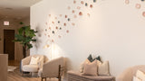 A modern interior lounge or waiting area with light wood flooring, cream upholstered armchairs, a ribbed bench with pillows, and a white wall decorated with many small circular wall pieces in muted pink, beige, and gray tones.