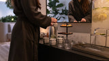 A woman in a dark brown robe stands at a beverage station, pouring from a metal kettle into glass cups. A large mirror reflects her, and a lemonade dispenser is visible on the right.