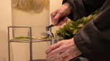 A person in a dark jacket pours a clear glass of water from a large clear beverage dispenser filled with lemon slices and green leaves, set on a dark table.