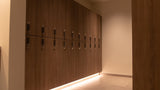 A modern hallway featuring a long wall of dark wood storage cabinets with integrated lockers and keys, illuminated by warm under-cabinet LED lighting.