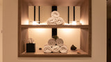 A built-in wooden wall shelf with warm lighting, styled with neatly rolled white towels, a tissue box, and small decorative objects against a beige wall.