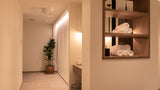 A warm, modern spa or bathroom interior with beige walls, a potted green plant in a hallway, and a built-in wooden shelf on the right holding neatly rolled white towels under warm lighting.