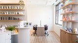 A bright, modern spa or beauty salon interior with light wood flooring, white and wood shelving filled with products, a central white pedestal counter, and two treatment chairs in the distance.