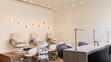 A modern spa or salon interior with four beige reclining chairs along a wall, white treatment tables with black lamps, rolling stools, and warm wall lights on a light wood floor.