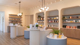 A bright, modern retail interior with curved white and blue display tables, built-in shelving, wood flooring, and multiple hanging pendant lights. No people or animals are present, and the scene appears to show the store layout rather than a single isolated product.
