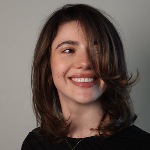 A smiling young woman with shoulder-length brown hair and bangs, wearing a black top, photographed against a plain light gray background.