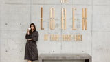A woman in a dark brown robe stands next to a concrete bench in front of a concrete wall with large gold letters reading 'THE LOREM AT LADY BIRD LAKE'.
